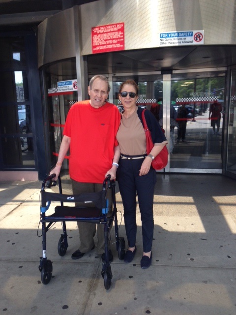 A man and woman stand outside some doors. The man has a walker.