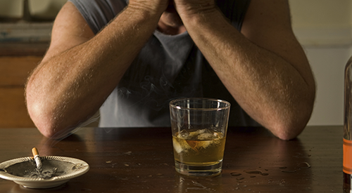 A man at a table covering his face with a cigarette and scotch in front of him.