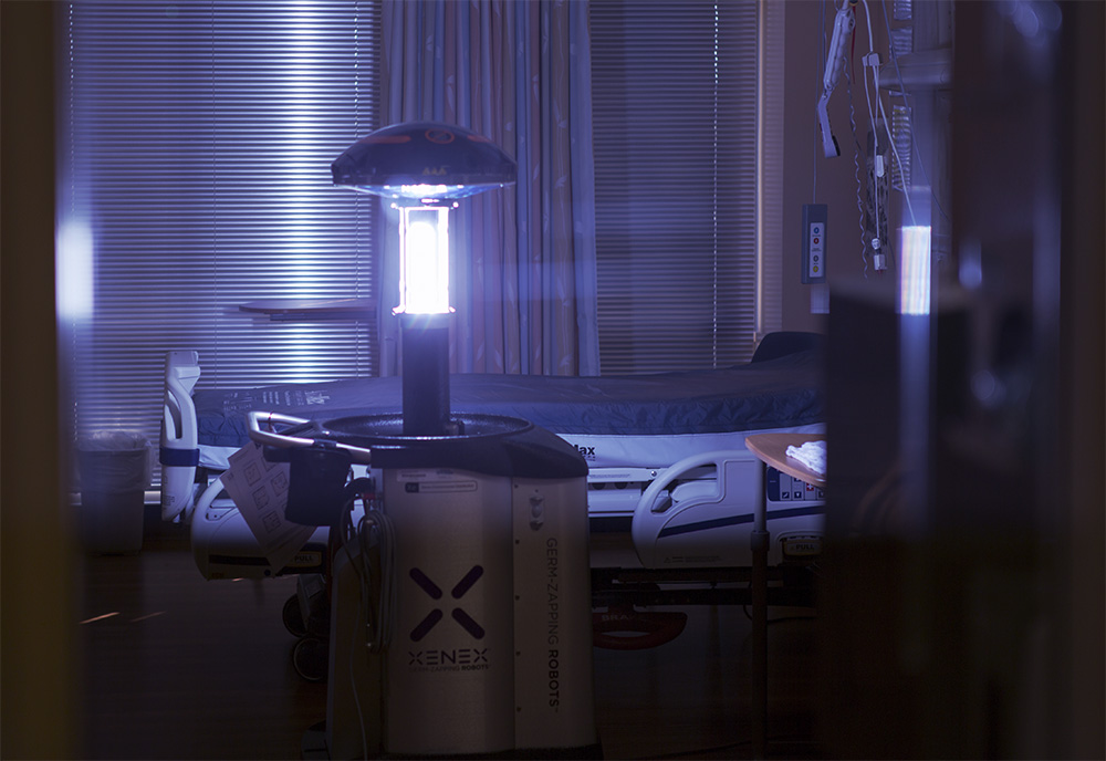 A tall, cylindrical machine stands in the center of a hospital room and emits blue light