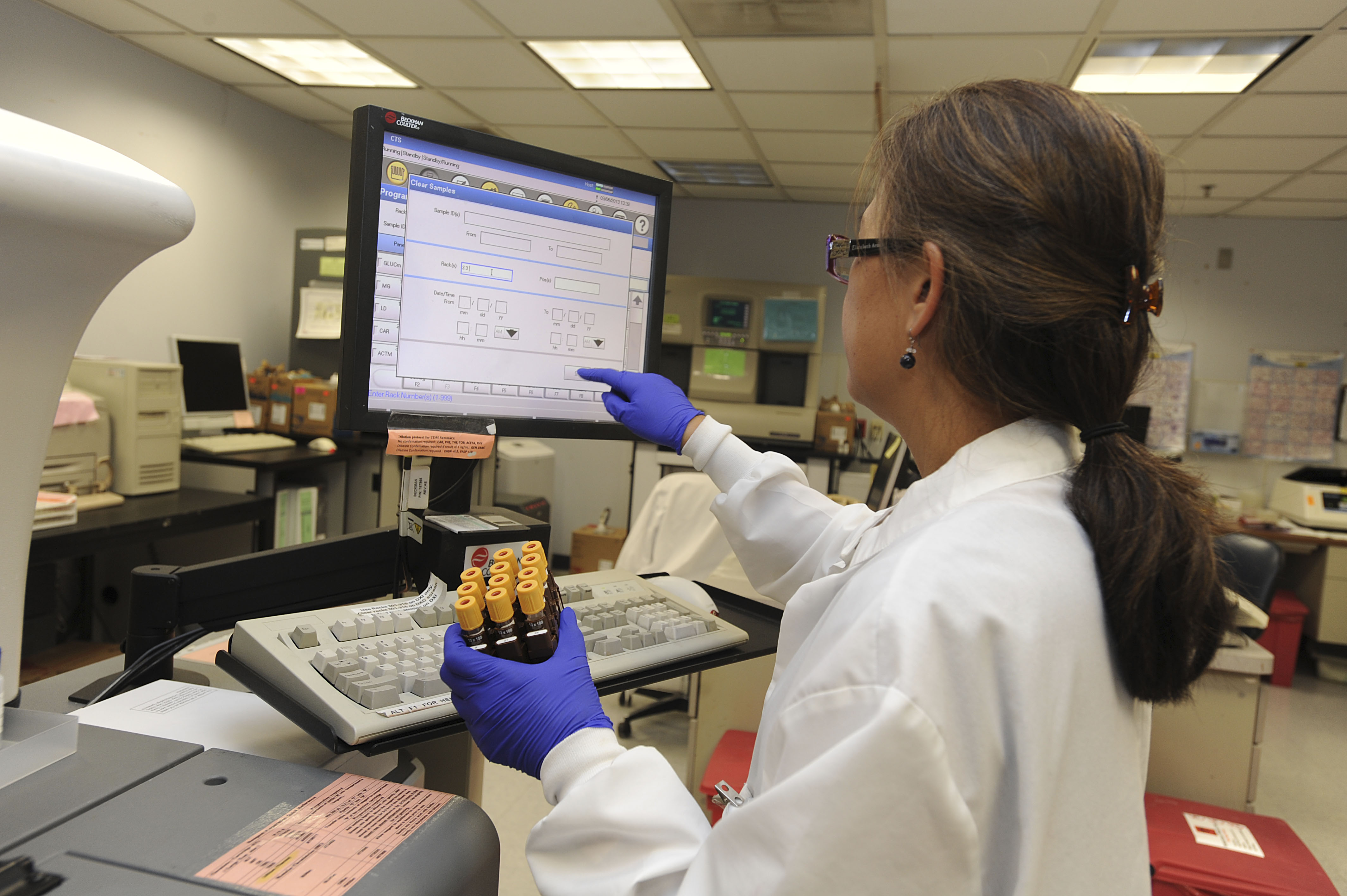 A woman in a lab coat holds viles while touching a computer screen.