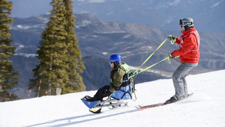 Two people ski, one in a seated skier