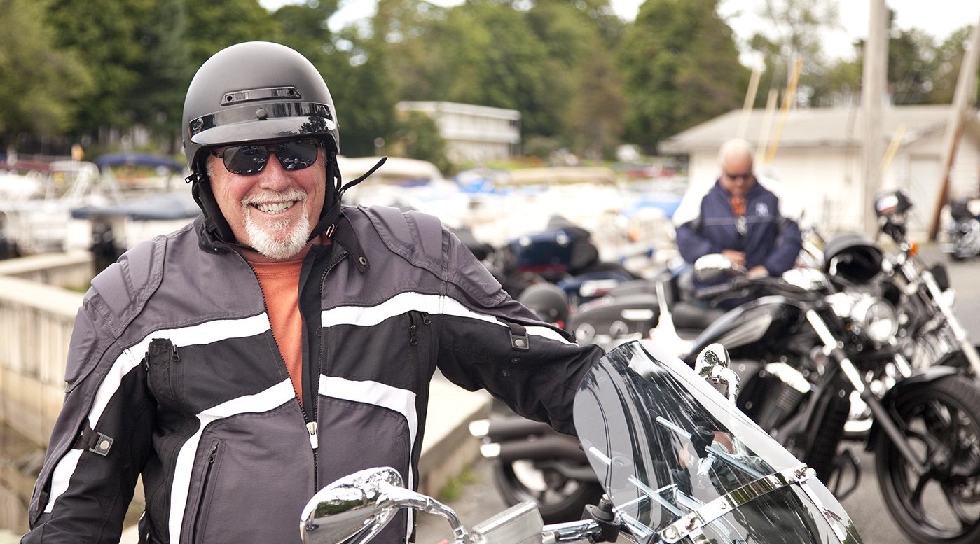 Motorcyclist smiling standing next to motorcycle on a cool riding day. Dressed in a warm jacket and helmet wearing sun glasses and parked in front of other motorcycles with unidentifiable biker behind him. Photo taken with Canon 5D Mark2 at 100 ISO, 24-115mm lens.