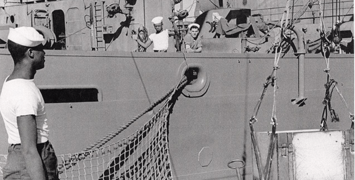 Photograph. A sailor looks onto a docked ship that is receiving supplies and munitions. Two sailors look toward the camera from the ship.