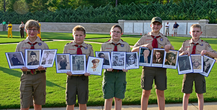 Five young boy scouts holding pictures of Veterans in front of a open grass field.