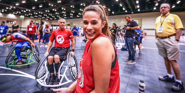 Woman smiling, sitting in a wheelchair on a basketball court with others in wheelchairs around her