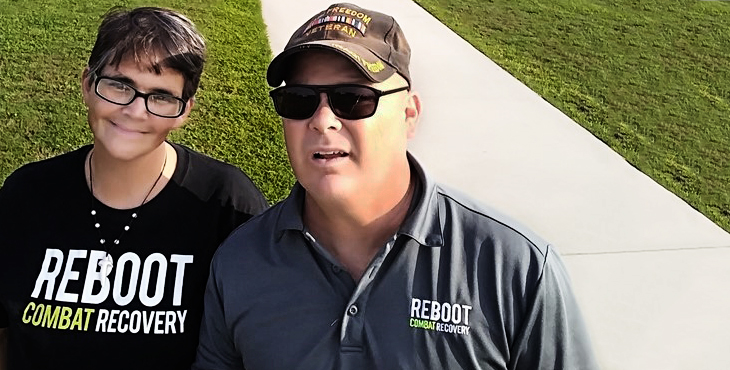 Picture shows two Veterans wearing "REBOOT COMBAT RECOVERY" apparel, looking at the camera with green grass and a sidewalk behind them.