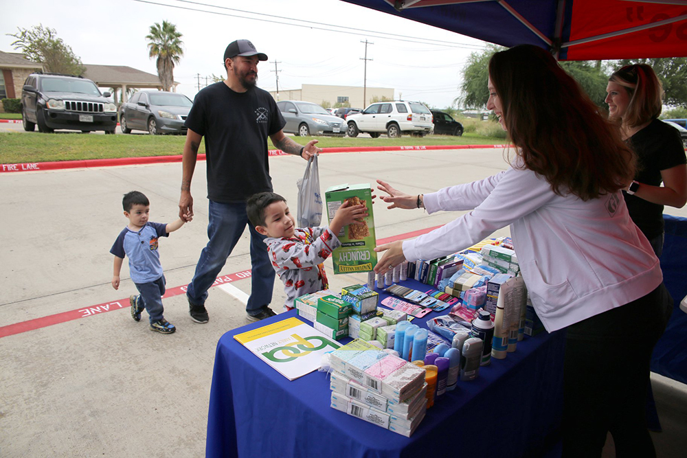 (Far right) Gina Guell smiles as fellow military spouse and volunteer, Lisa Quirk, reaches out to accept a large box of granola bars being donated by four-year-old Ivan Hernandez and his family, during the Boots & Badges care package drive held October 26, 2018, at Vet Center in Laredo, Texas. Local resident and businessman Hector Hernandez said he brought his sons Ivan and Jack [age 2] in order to set a good example for his children from an early age about the importance of supporting our nation's Veterans and giving back to your community.