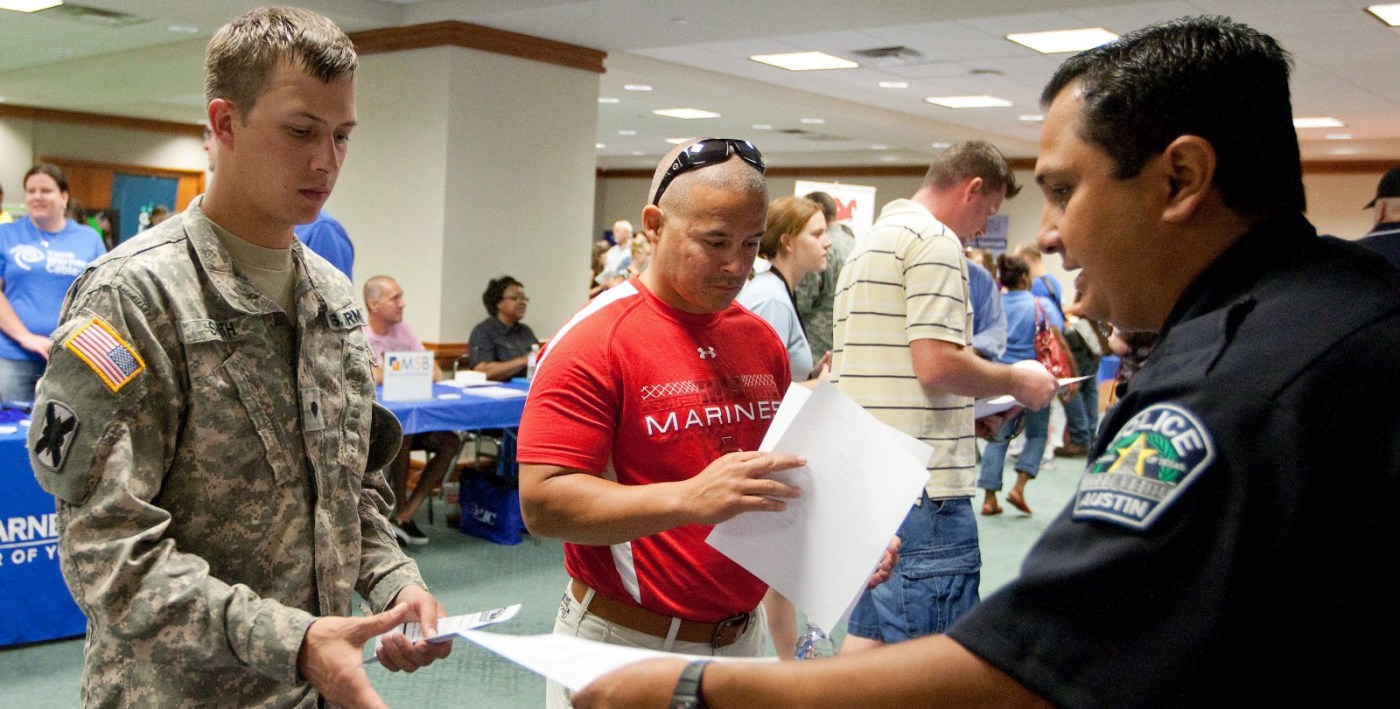 Picture shows a police officer handing a piece of paper to a service member in uniform. Other people, including Veterans in the background.