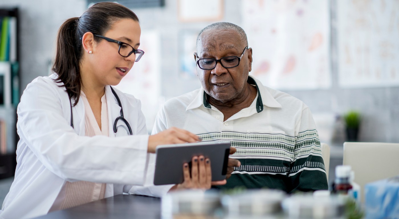 Older patient in a health care office receives medical counsel from a practitioner holding a tablet device.