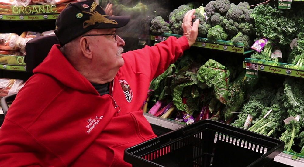 A male Veteran in a red sweatshirt reaches to place broccoli in a shopping cart in a supermarket