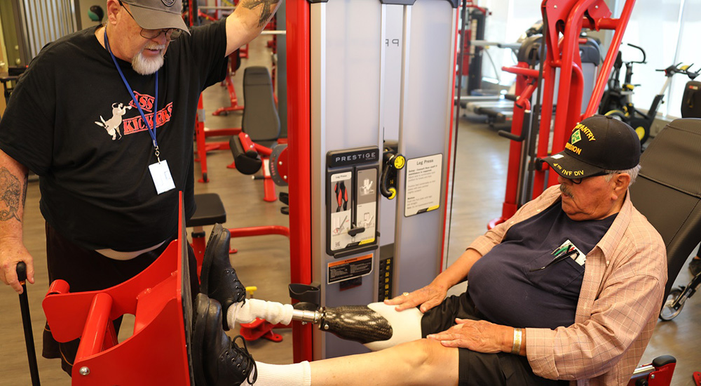 One man stands next to a piece of gym equipment while another man, who has a prosthetic leg, does leg presses.