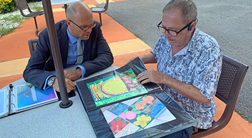 Two men sitting at an outdoor table looking at a portfolio of paintings