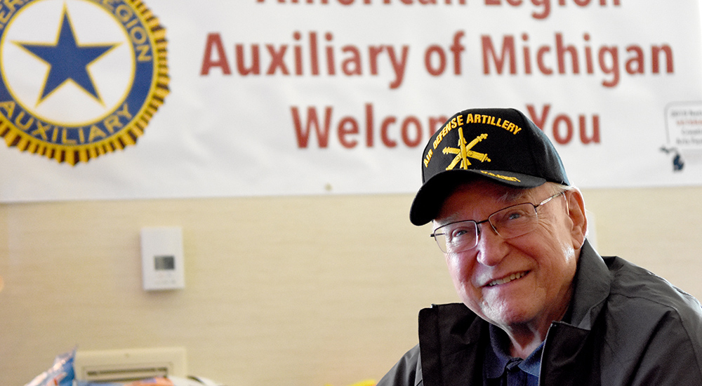 Man wearing an Air Defense Artillery cap sits in front of a banner that reads American Legion Auxiliary of Michigan Welcomes You at the National Veterans Creative Arts Festival
