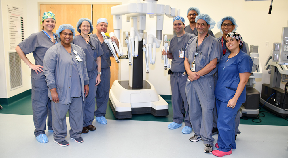 Nine members of a surgical team stand next to a robotic system