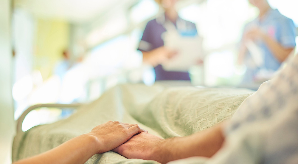 A hospital visitor's hand holds a patient's hand in bed of a hospital ward. In the blurred background a young nurse is chatting to the ward sister about the patient's care.