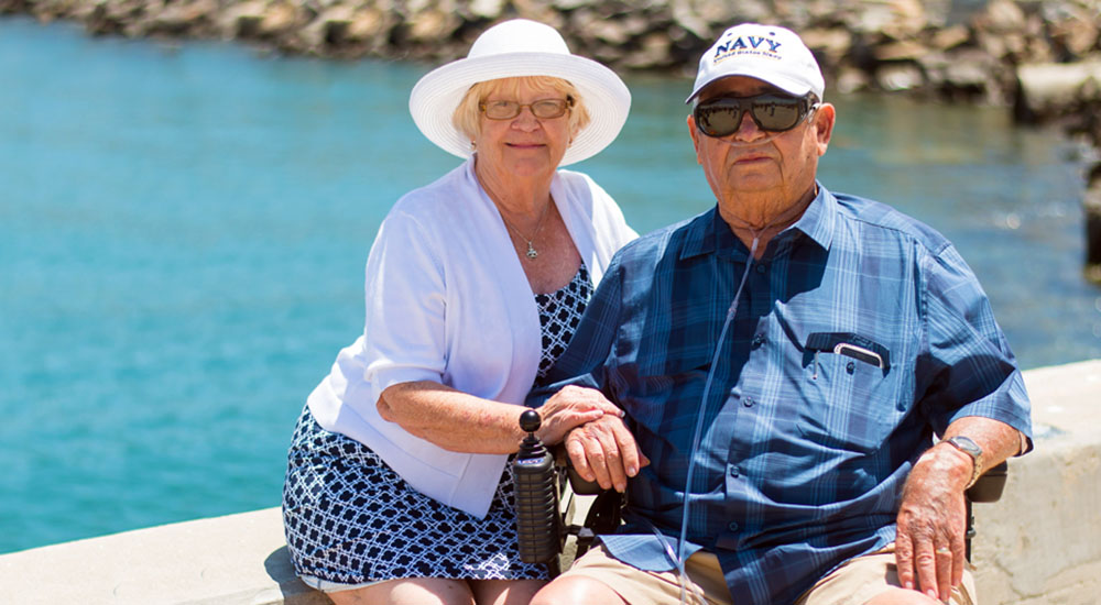 And elderly man wearing a Navy Veteran cap and his caregiver wife sit by the water