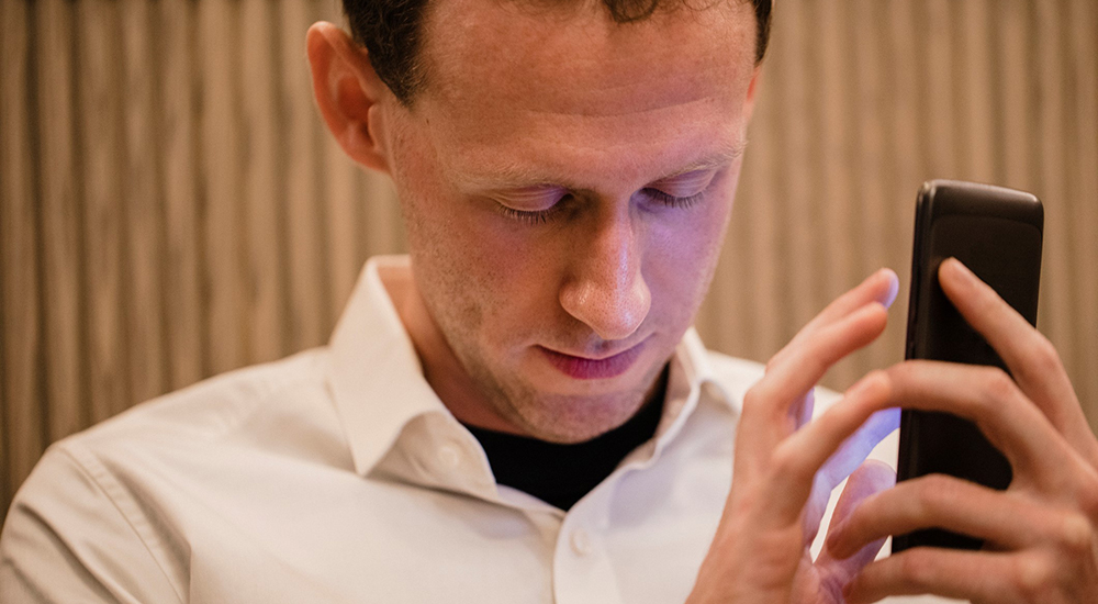 A close-up of a man wearing casual clothing, he has his smartphone in his hand and he is using a visually impaired mobile app to help assist him.