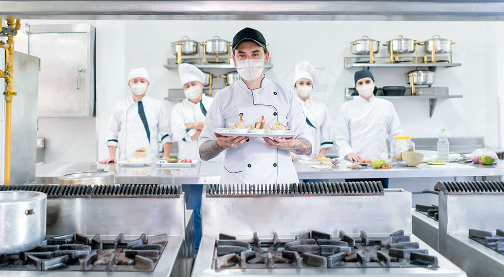 Cook with a catering team wearing facemasks in a commercial kitchen while presenting a plate and looking at the camera â pandemic lifestyle concepts
