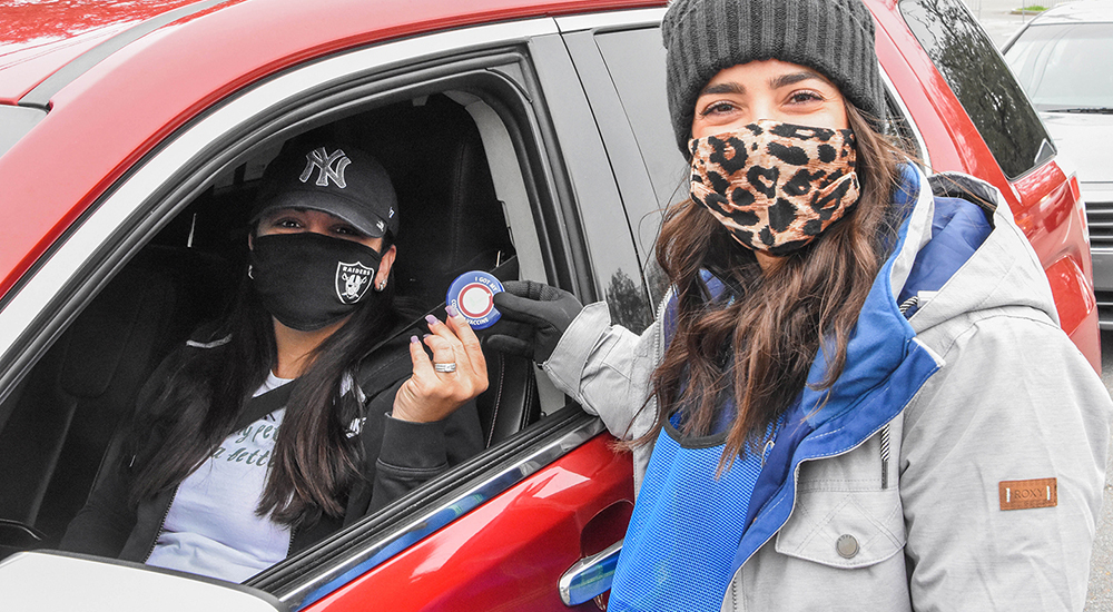 Woman in mask hands badge to woman in car wearing mask