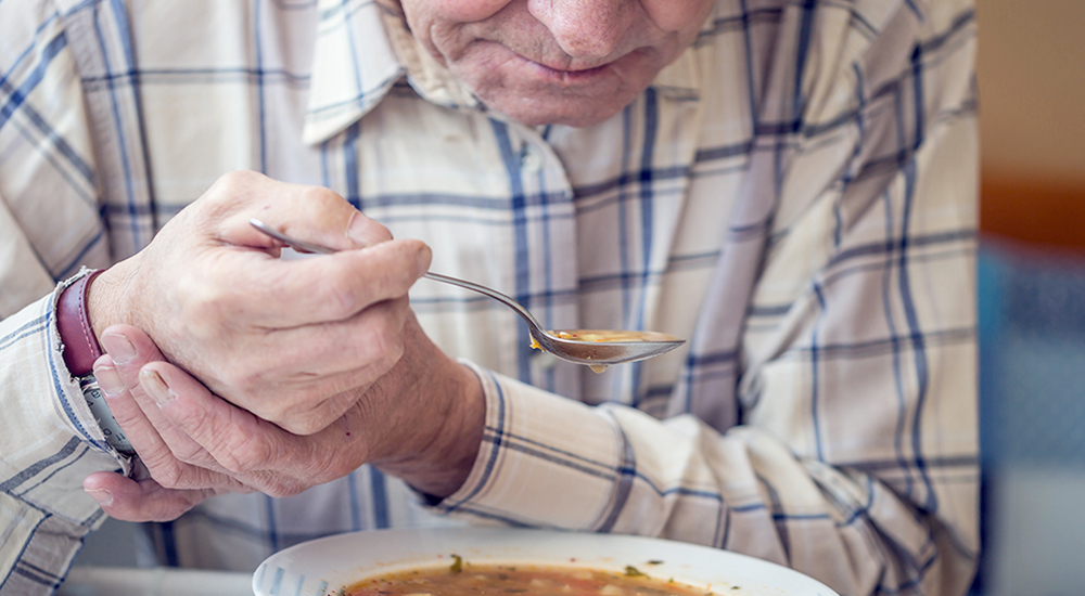 Elderly man with holds spoon in both hands as he eats soup.