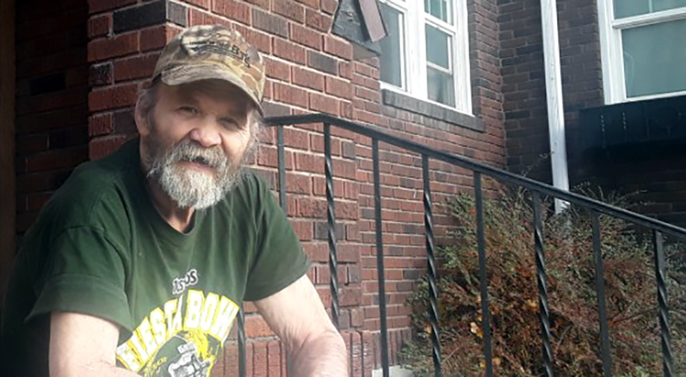 Bearded Veteran in fatigue cap sitting on the porch of a home he found through HUD-VASH