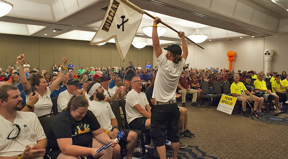 Man raising flag to large National Veterans Summer Sports Clinic audience