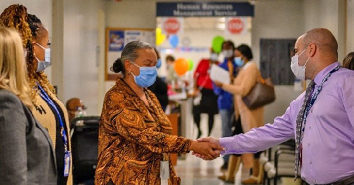 Two people wearing masks shake hands in a hallway. 