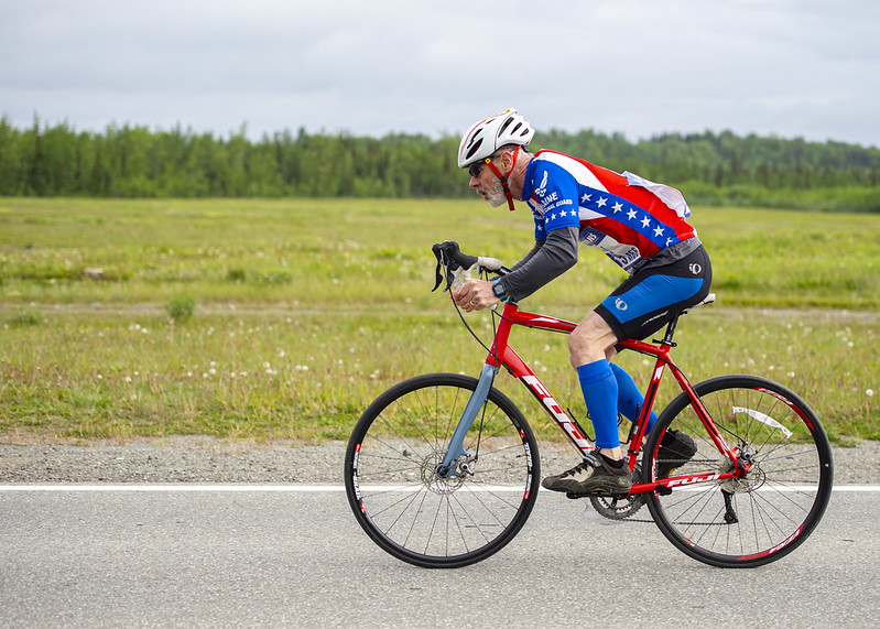 Veteran wearing red, white and blue patriotic bicycle gear, riding a bicycle along a roadway.