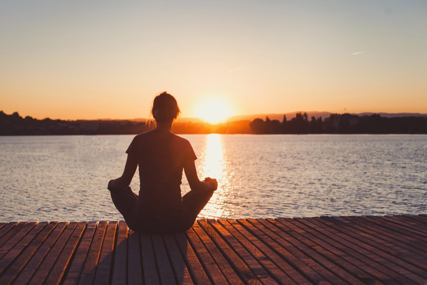 Woman sitting in a yoga sit, meditating on a boat dock while the sun sets over the water.