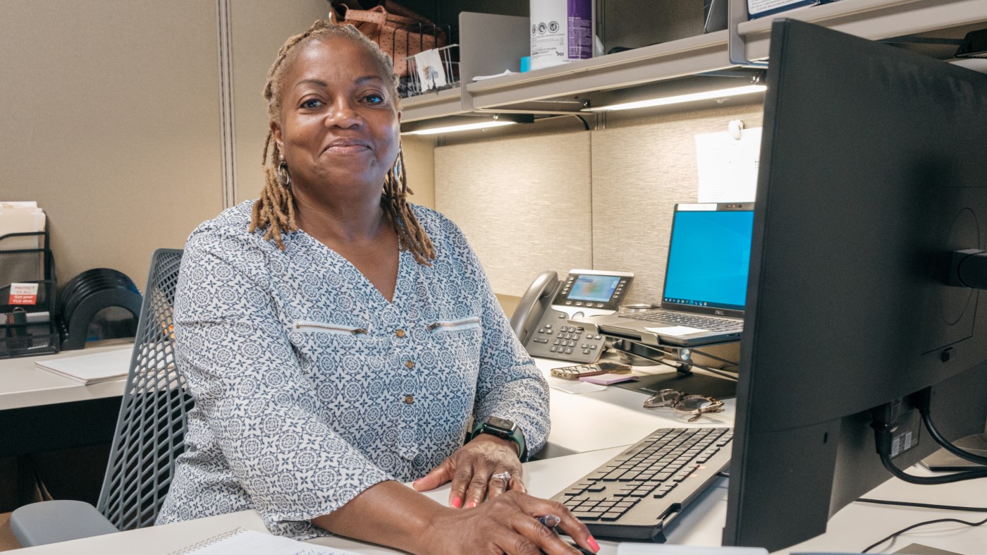 A female VA recruiter sits at her computer, smiling toward the camera, ready to discuss the application process.