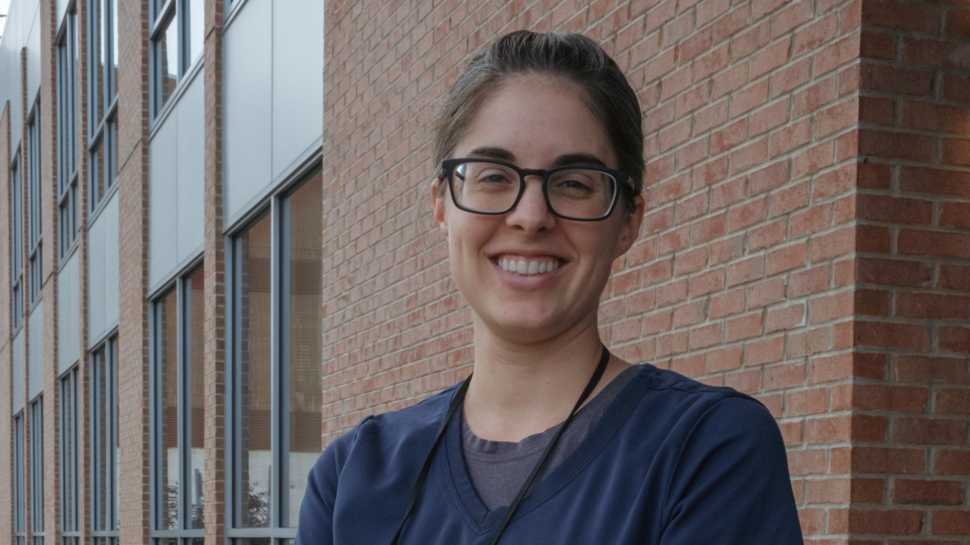 A smiling woman wearing glasses and scrubs outside a VA facility.