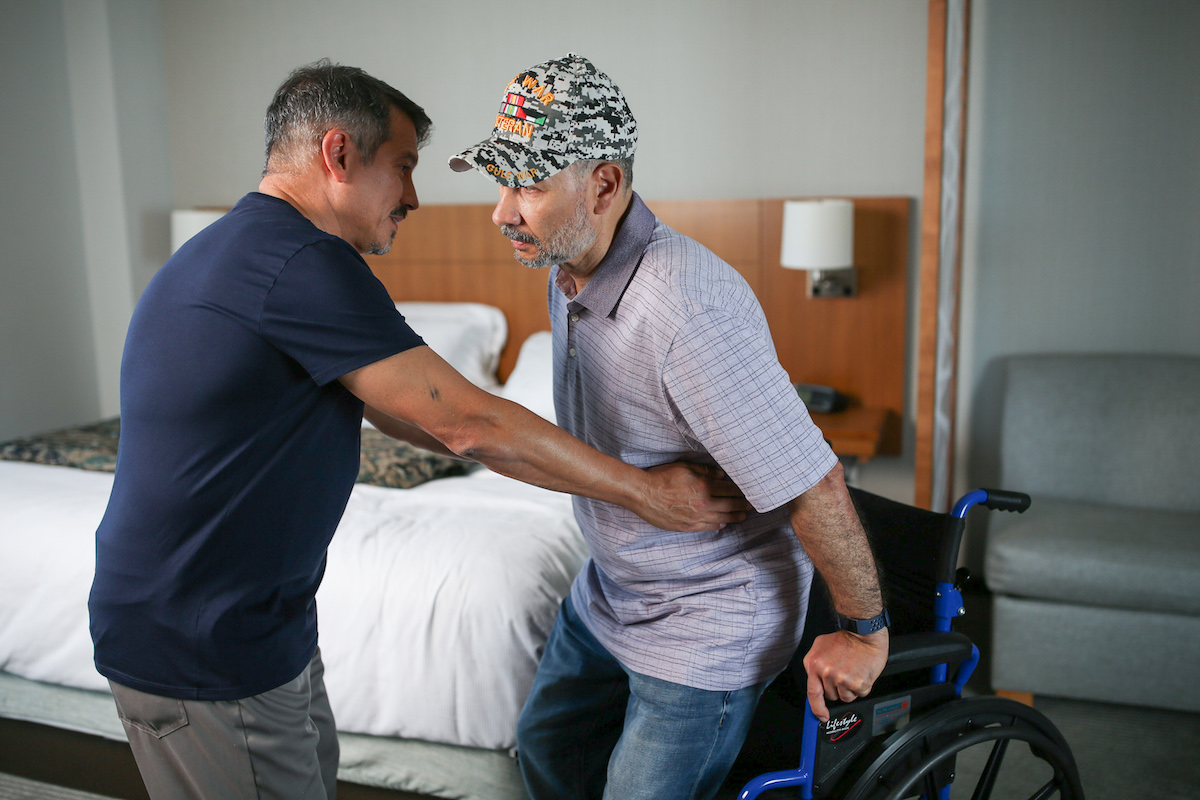 Man assisting other man up from a wheelchair.